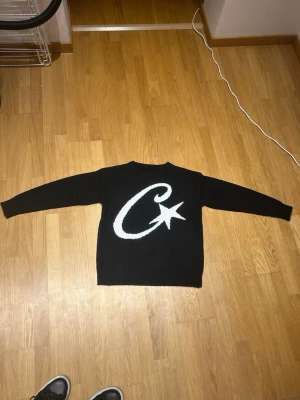 Svart stickad tröja med stjärntryck/ corteiz - Svart stickad tröja med lång ärm och rund hals. På framsidan finns en stor vit design med en böjd linje och en stjärna. Tröjan har en klassisk passform och är perfekt för kyligare dagar. 