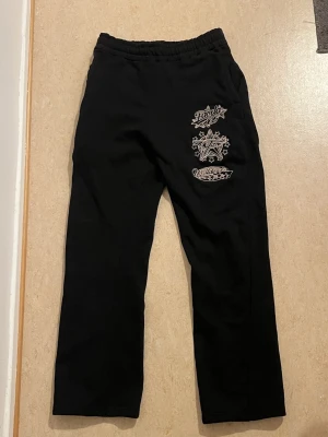 Svarta mjukisbyxor från Favela Clothing - Svarta mjukisbyxor med elastisk midja och coola vita tryck på ena benet. Byxorna har raka ben och en bakficka. Perfekta för chill dagar och har en avslappnad passform. Materialet känns mjukt och bekvämt mot huden.