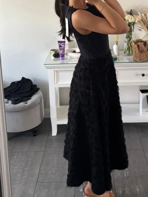 Svart långklänning med struktur - Säljer en svart långklänning med ärmlös överdel i slät textil och kjol i strukturerat material som ger en lyxig känsla. Klänningen har en markerad midja och elegant siluett.  Dragkedja i sidan för enkel påklädning..Ny med prislappen kvar