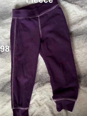 Mörklila fleecebyxor med muddar - Mörklila fleecebyxor med bred resår i midjan och mjuka muddar vid bensluten. Perfekta för kalla dagar och superbekväma att ha på sig. Snygga kontrastsömmar ger en sportig vibe.🌸 Fint använt skick. Storlek 98