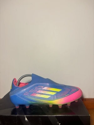 Adidas F50 blå/rosa fotbollsskor - Säljer ett par Adidas F50 fotbollsskor i en riktigt fet färgkombo av blått, rosa och gult. Skorna har en strömlinjeformad design utan snörning, med syntetmaterial och tydliga F50-detaljer på sidan och sulan. Perfekta för dig som vill sticka ut på planen.