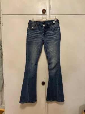 Blå bootcut jeans från True Religion - Säljer ett par blå bootcut jeans från True Religion med snygga slitningar och kontrastsömmar. Jeansen har jag använt 10-15 ggr.Materialet är jeans i bomull och passformen är normal. Är i storlek 26