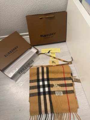 Rutig beige cashmere halsduk Burberry - Exklusiv halsduk från Burberry i klassiskt rutigt mönster med beige, svart, vit och röd. Tillverkad i 100% kashmir och har broderad Burberry-logga. Mjuk och lyxig känsla, perfekt accessoar för att lyfta din outfit.                  Storlek 230 cm X 30 cm. Väldigt skön nu inför kalla dagar :)