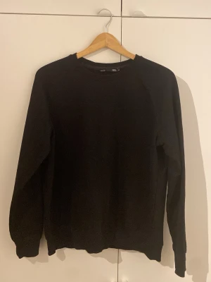 Svart sweatshirt från 157 - En enkel och stilren svart sweatshirt från 157. Tröjan har rund halsringning och långa ärmar. Tillverkad i mjuk bomull som gör den skön att bära. Perfekt basplagg att matcha med jeans eller kjol.