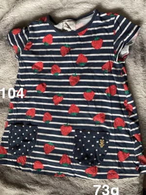 Randig tunika med jordgubbar och prickiga fickor - Söt topp från Holly & whyte i marinblått och vitt med horisontella ränder och röda jordgubbar över hela. Två mörkblå fickor framtill med vita prickar och ett litet broderat märke. Kortärmad och rund halsringning, perfekt för lekfulla dagar.🌸 Fint använt skick. Storlek 104