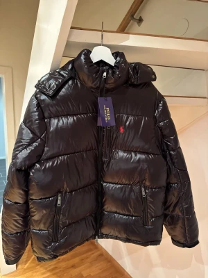 Polo Ralph lauren vinterjacka - Snygg och exklusiv dunjacka från Polo Ralph Lauren i klassisk svart med glansig finish. Jackan har en modern, sportig design och är både varm och lätt – perfekt för höst och vinter.
