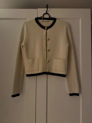 Beige kofta från Zara med svarta detaljer - Säljer en beige stickad kofta från Zara med svarta ribbade kanter vid hals. Koftan har guldfärgade knappar framtill och två fickor. Stilig och passar till många tillfällen. Aldrig använd.🤍