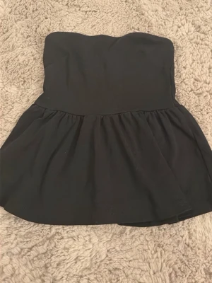 Svart bandeau topp från Gina Tricot - Säljer en svart bandeau topp från Gina Tricot i storlek XS. Toppen är axelbandslös med en utsvängd, lätt volang nedtill och har en enkel, stilren design. Perfekt att styla med jeans eller kjol för en trendig look.