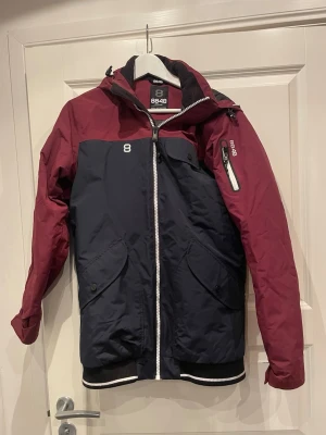 8848 Altitude marinblå och vinröd jacka - Säljer en snygg jacka från 8848 Altitude i marinblått och vinrött. Jackan har huva, dragkedja framtill, fickor med lock och en extra ficka på ärmen. Sportig look med ribbade muddar och vita detaljer. Perfekt för kyliga dagar.