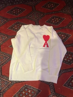 Vit stickad tröja Ami Paris  - Säljer en vit stickad tröja från Ami Paris med röd hjärtdetalj och bokstaven A på bröstet. Tröjan har rund halsringning, långa ärmar och ribbade muddar vid ärmslut och nederkant. Perfekt för dig som gillar stilrena plagg med en twist.