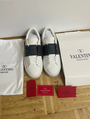 Valentino Garavani Open sneakers vit/mörk blå  - Säljer ett par Valentino Garavani Open sneakers i vitt skinn med bred mörk blå  rem över vristen. Klassisk rund tå, vita snören och subtila nitar på hälen. Snygg och stilren design som passar till allt. Kommer med originalkart firmapåse och certifikat. Pris kan diskuteras 