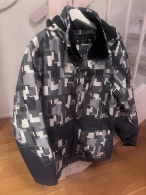 Grå mönstrad vindjacka med huva - Säljer en grå och vit vindjacka från Tenson med grafiskt rutigt mönster. Jackan har huva, dragkedja och stora fickor framtill. Perfekt för dig som gillar streetwear och vill sticka ut med en unik look.