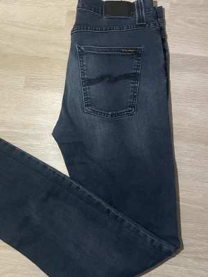Mörkblå jeans från Nudie Jeans, W32/L34 - Säljer ett par mörkblå jeans från Nudie Jeans med klassisk femficksdesign och diskret slitning på framsidan. Jeansen har raka ben och normal passform, tillverkade i mjukt bomullsmaterial. Perfekta för dig som gillar stilrena och tidlösa jeans.