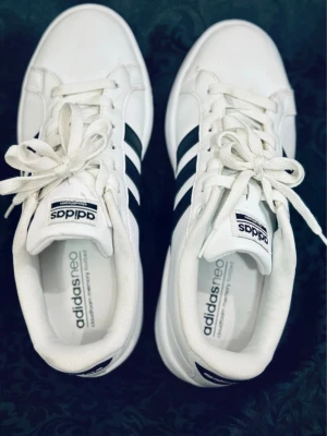 Vita Adidas Neo sneakers med svarta ränder - Säljer ett par fräscha vita Adidas Neo sneakers med klassiska svarta ränder på sidorna och svart Adidas-logga på hälen. Skorna har snörning, rund tå och är tillverkade i syntetmaterial med memory foam innersula för extra komfort.