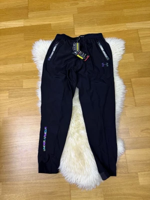 Svart Under Armour byxor, HELT NY!!! - Helt ny!!! Snygga svarta träningsbyxor från Under Armour med elastisk midja och snörning. Byxorna har dragkedjeförsedda fickor och coola regnbågsskimrande loggor på benet och vid fickorna. Tillverkade i lätt och funktionellt syntetmaterial, perfekta för träning.