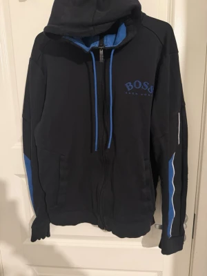 Svarta mjukisbyxor från Hugo Boss - Svarta mjukisbyxor från Hugo Boss med blå detaljer och logga på benet. Byxorna har resår och snörning i midjan samt blå sidopaneler. Perfekta för en chill och sportig look.