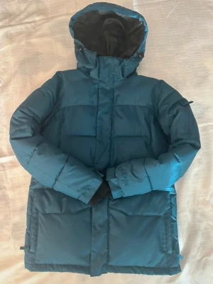 Blå pufferjacka från Tribute - Säljer en blå pufferjacka från Tribute med huva och stora fickor. Jackan har dragkedja, ribbade muddar vid ärmsluten och broderad logga på ärmen. Insidan har en praktisk innerficka med dragkedja. Perfekt för kalla dagar!