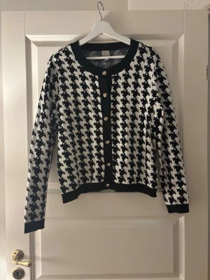 Svartvit rutig kofta från H&M - Säljer en svartvit kofta från H&M i storlek M med klassiskt rutigt mönster. Koftan har guldfärgade knappar framtill, rundad halsringning och ribbade muddar. Perfekt för dig som vill ha en statement piece i garderoben.