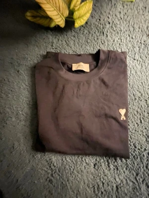 Svart t-shirt från AMI Paris - Svart t-shirt från AMI Paris med klassisk passform och korta ärmar. På bröstet finns ett broderat hjärta med bokstaven A i beige. Tillverkad i mjuk bomull och har rund halsringning. Diskret AMI-broderi i nacken.