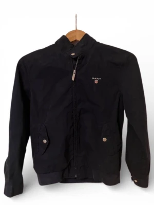 Svart vindjacka från GANT i SS - Klassisk svart vindjacka från GANT med ståkrage, dragkedja framtill och två fickor med knappstängning. Jackan har diskret GANT-logga och emblem på bröstet samt ribbade muddar vid ärmslut och nederkant. Perfekt för dig som gillar stilren och enkel design.