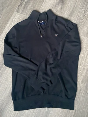 Halv zip GANT - Klassisk svart långärmad tröja från GANT med half zip och liten broderad logga på bröstet. Tröjan har ribbade muddar och hög krage, perfekt för lager på lager. Tillverkad i högkvalitativ merino ull för skön känsla.