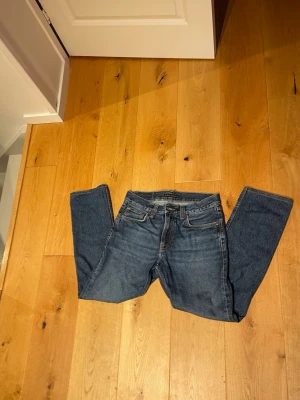 Mörkblå raka jeans från Nudie - Säljer ett par klassiska mörkblå jeans från Nudie med raka ben och fem fickor. Jeansen har normal passform och är tillverkade i slitstark denim med snygga kontrastsömmar och Nudie-logga på bakfickan.