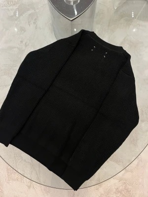 Svart stickad tröja Maison Margiela storlek m - Storlek M. Säljer en svart stickad tröja från Maison Margiela med ribbad struktur och rund hals. Tröjan har långa ärmar och klassisk passform. Diskreta vita stygn på ryggen ger en unik touch. Perfekt för dig som gillar stilrena och coola plagg.