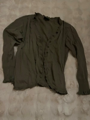 Olivgrön kofta från Wästgöta - Säljer en olivgrön kofta från Wästgöta by Cellbes i storlek 42/44. Koftan har långa ärmar, volangkant längs öppningen och nederkanten samt knappar framtill. Perfekt för lager på lager och enkel att styla till olika outfits. Kan användas som bara tröja ochså