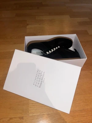 Maison Margiela svarta sneakers - Maison Margiela sneakers i svart skinn och mocka med vita snören och klassisk gummisula. Snygg minimalistisk design med rund tå och diskreta paneler. Kommer i originalkartong. Perfekta för dig som gillar stilrena och exklusiva sneakers.