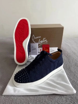 Mörkblå sneakers från Christian Louboutin - Säljer ett par mörkblå sneakers från Christian Louboutin med klassisk röd sula och coola nitar på tån. Skorna har snörning och är tillverkade i mocka med vita platta sulor. Perfekta för dig som vill sticka ut med exklusiv design och edgy detaljer.