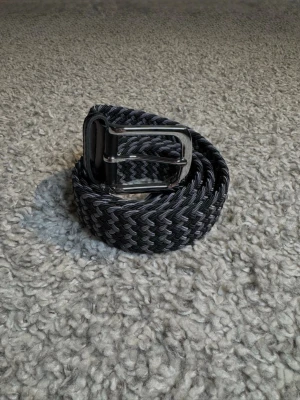 Svartgrått Flätat Bälte - Svart-grått flätat bälte med metall och läder spänne. Gjord av elastiskt material som anpassas precis efter behov. 105 cm lång