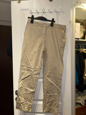 Beiga chinos från GANT - Säljer ett par klassiska beiga chinos från GANT. Byxorna har raka ben, bälteshällor och snygga bakfickor med lock. Tillverkade i mjuk bomull som är sköna att ha på sig hela dagen. Perfekta för en clean och stilren look. Lite mindre i storlek!