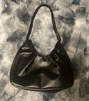 Svart axelväska i skinnimitation - Säljer en stilren svart axelväska i skinnimitation med mjuk, slät yta och enkel design. Väskan har en bred axelrem och rymligt huvudfack, perfekt för att bära med sig det viktigaste. Passar dig som gillar minimalistisk och trendig stil.