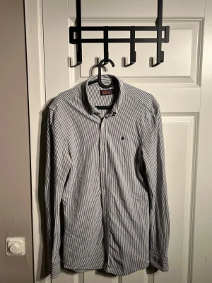 Blå och vit Randig skjorta från Morris - Snygg vit och marinblå randig skjorta från Morris med klassisk krage och knappar framtill. Skjortan har lång ärm och en liten broderad logga på bröstet. Mer frågor eller bilder är det bara att fråga!