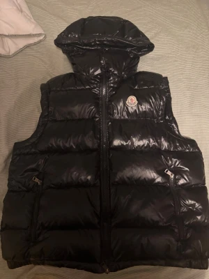 Svart dunväst från Moncler - Säljer en svart, glansig dunväst från Moncler med avtagbarhuva och två dragkedjeförsedda fickor. Köpte västen av en polare för 2 år sedan men har stannat på klädhängaren sen dess. Är osäker på om den är äkta eller inte då jag inte har så stor koll på moncler, därav priset. Storlek 4 men passar mig som har L, passa även M. NFC fungerar. Vid frågor så äre ba höra av sig. //mvh MB