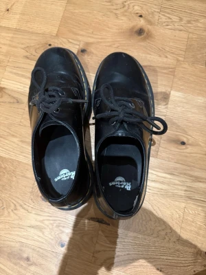 Svarta Dr. Martens 1461 i skinn - Klassiska Dr. Martens 1461 i svart skinn med robust, räfflad sula och gula sömmar. Skorna har rund tå, snörning och den ikoniska Dr. Martens-loggan på innersulan. Perfekta för dig som vill ha en tidlös och edgy look.