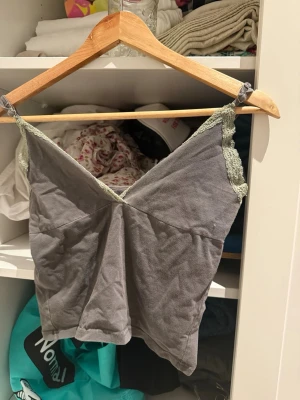 Grå linnetopp med spetskant - Säljer en grå linnetopp med tunna axelband och ljusgrön spetskant längs urringningen. Toppen har en v-ringad form och är tillverkad i mjuk bomull. Perfekt för varma dagar eller som lager under en skjorta.