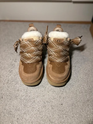 UGG beige sneakers med ullfoder - Säljer ett par snygga UGG sneakers i beige mocka och mesh med fluffigt vitt ullfoder. Skorna har grova, mönstrade snören i beige och vitt samt en rejäl sula för bra grepp. Perfekta för kalla dagar och riktigt sköna att ha på sig.