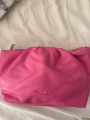 Elizabeth Arden väska - Säljer en snygg rosa kuvert/smink  från Elizabeth Arden till ett billigt pris!. Väskan är gjord i mjukt syntetmaterial och har en guldfärgad dragkedja. Den har en puffig, veckad design som ger en cool och modern look. Perfekt storlek för det viktigaste och enkel att bära med sig.