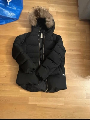 Svart pufferjacka från Lexington XS - Säljer en svart pufferjacka från Lexington i storlek XS. Jackan har huva med fuskpäls, dragkedja framtill och ett diskret Lexington-märke på ärmen. Perfekt för kalla dagar och riktigt skön att bära.