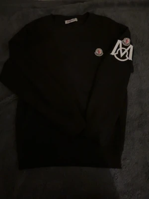 Moncler tröja  - Säljer en svart sweatshirt från Moncler med klassisk rund hals och stora vita Moncler-loggor på ärmen samt bröstet. Tröjan har lång ärm och är tillverkad i mjuk bomull. Perfekt för dig som gillar stilrena och exklusiva plagg.