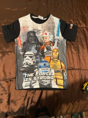 LEGO Star Wars t-shirt svart - Svart t-shirt med färgglatt LEGO Star Wars-tryck framtill. Motiv med Darth Vader, Stormtrooper, R2-D2, C-3PO och Luke Skywalker i LEGO-version. Korta ärmar, rund hals och tryck på ena ärmen. Framsida i polyester, rygg och ärmar i bomull.