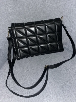 Svart quiltad axelväska i skinnimitation - Säljer en svart quiltad axelväska med justerbar rem. Väskan har ett geometriskt mönster på framsidan och flera fack inuti för bra organisering. Tillverkad i skinnimitation med en stilren look som passar till många outfits. 25 cm bred och allt får plats!!