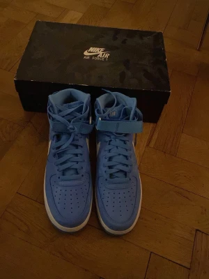 Nike Air Force 1 Hi Retro University Blue - Säljer ett par Nike Air Force 1 Hi Retro QS i färgen University Blue/Summit White. Höga sneakers med klassisk snörning, kardborreband vid ankeln och vit sula. Tillverkade i skinn med perforerad tå och ikonisk Nike-logga på hälen.