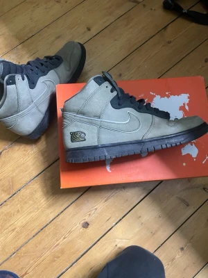 Nike Dunk High SB x Souldgoods - Unika Nike Dunk High SB med olivgröna mockapaneler, svarta snören och svart sula. Sidan har en broderad tiger och plösen har texten 'To The World From Beijing'. Perfekta för dig som gillar streetwear och vill sticka ut.