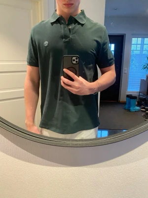 Mörkgrön piké från Timberland - Säljer en mörkgrön pikétröja från Timberland i regular fit. Tröjan har klassisk krage, två knappar och en liten broderad Timberland-logga på bröstet. Tillverkad i 100% ekologisk bomull för en skön och avslappnad stil.