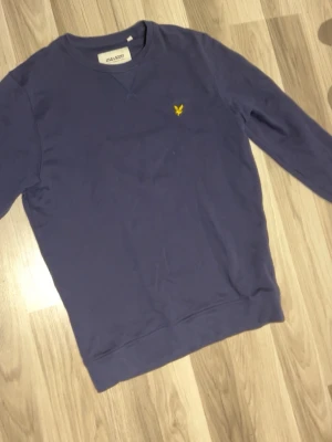 Marinblå sweatshirt från Lyle & Scott - Säljer en marinblå sweatshirt från Lyle & Scott i ekologisk bomull. Tröjan har rund hals, lång ärm och den klassiska gula loggan broderad på bröstet. Perfekt för dig som gillar stilrena och bekväma plagg.