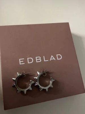 Peak hoops Edblad  - Coola örhängen i silverfärg från Edblad med chunky design och spetsiga nitar runt hela ringen. Perfekta för dig som vill ha en edgy vibe till din outfit. Metallfinishen ger ett stilrent och modernt intryck.