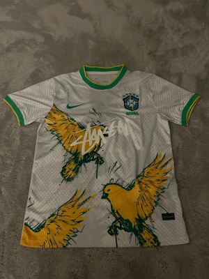 Nike x Stüssy Brasil fotbollströja - Hör av innan köp! Unik vit Nike fotbollströja i samarbete med Stüssy och Brasil, med gröna och gula detaljer. Framsidan har stora grafiska illustrationer av gula fåglar och Stüssy-loggan tryckt över bröstet. Kragen och ärmsluten har gröna och gula ränder. Perfekt för dig som vill sticka ut på planen.