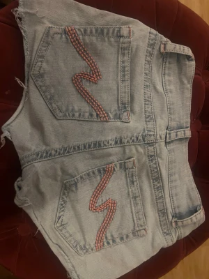 Ljusblå jeansshorts från Nelly - Säljer ett par ljusblå jeansshorts från Nelly med  fransiga benslut. Shortsen har bakfickor med cool rosa broderad detalj och dubbla knappar framtill. Perfekta för sommaren och riktigt snygga till sneakers eller sandaler. Finns kanin i hemmet.